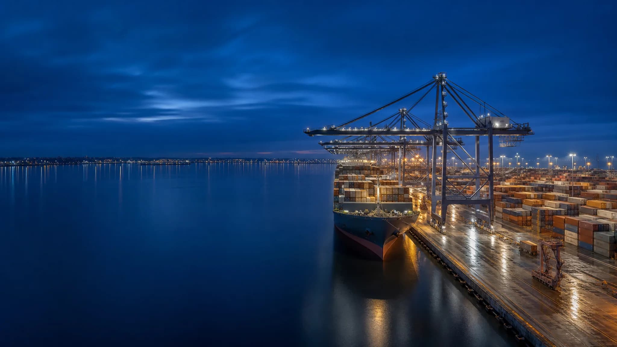 A container terminal at blue hour, with ship-to-shore gantry cranes over a moored container vessel