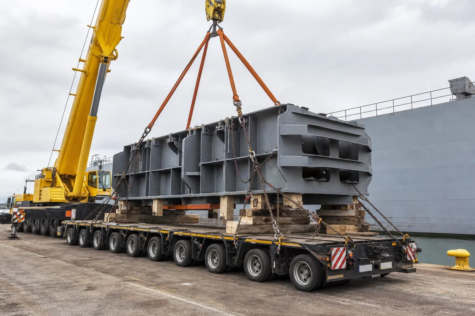 An oversized industrial component on a multi-axle flatbed trailer being lifted by a mobile crane at a quayside